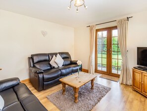 A living room with a black leather sofa, a wooden coffee table, a television on a wooden cabinet, and a large window with curtains. - Wild Duck Lodge, Mawdesley (Ormskirk)