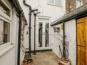 A narrow alleyway with a white door, black pipes, and two potted plants.