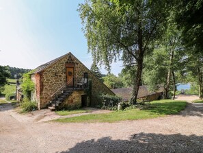 A stone building with a staircase leading to a door, surrounded by greenery and a gravel path.