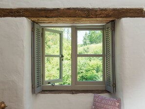 Ein Fenster mit Rollläden, Blick ins Grüne und ein Kissen mit Schriftzug an der Wand.