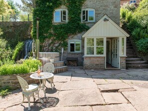 Ein Steinhaus mit einer kleinen Gartenlaube, einer hölzernen Bank und einer Glastür, die in einen überdachten Bereich führt.