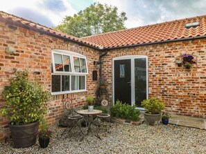 A brick house with a tiled roof, a small patio with a table and chairs, and potted plants. - The Barn at Orchard Farm, York (York)