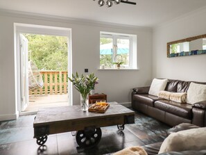 Un salon avec un canapé en cuir marron, une table basse en bois sur laquelle se trouve un plateau avec de la nourriture et des boissons, et une vue sur une terrasse en bois, accessible par une porte-fenêtre coulissante.