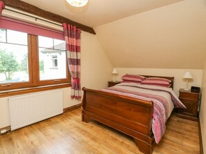 Un dormitorio con cama de madera, ventana con cortinas, mesita de noche con lámpara y radiador.