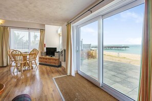 Une pièce dotée d’un plancher en bois, d’une table à manger avec des chaises, d’un téléviseur à écran plat et d’une grande porte-fenêtre coulissante donnant sur une plage avec une jetée.