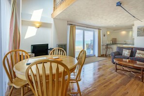 Une pièce comprenant une table à manger en bois, des chaises, un téléviseur, un canapé et une vue sur la plage, visible à travers une grande fenêtre.