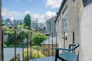 A stone building with a staircase leading to a balcony, surrounded by greenery and other stone buildings.