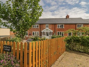 Een huis van baksteen met een veranda, een houten hek en een bord waarop staat 'Mulberry Cottage'.