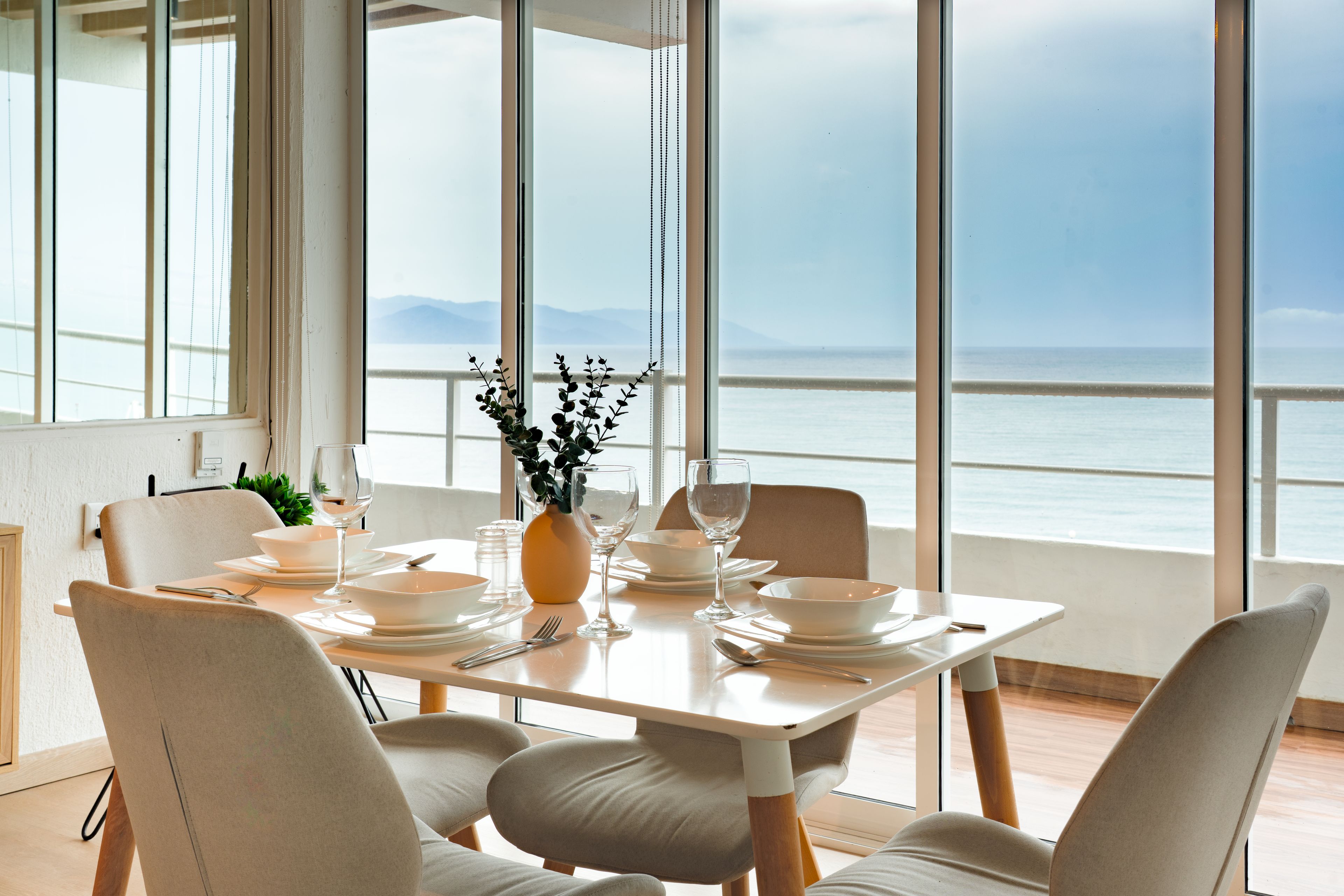 Une table à manger est dressée avec de l’assiette, des bols et des verres blancs, et est placée dans une pièce dotée de grandes fenêtres donnant sur un plan d’eau.
