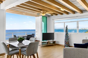 Un salon moderne avec un coin repas, de grandes fenêtres offrant une vue sur la mer, un téléviseur et un sapin de Noël décoré.