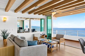 A modern living room with a sofa, coffee table, and chairs, a dining area with a table and chairs, and a large window with a view of the sea.