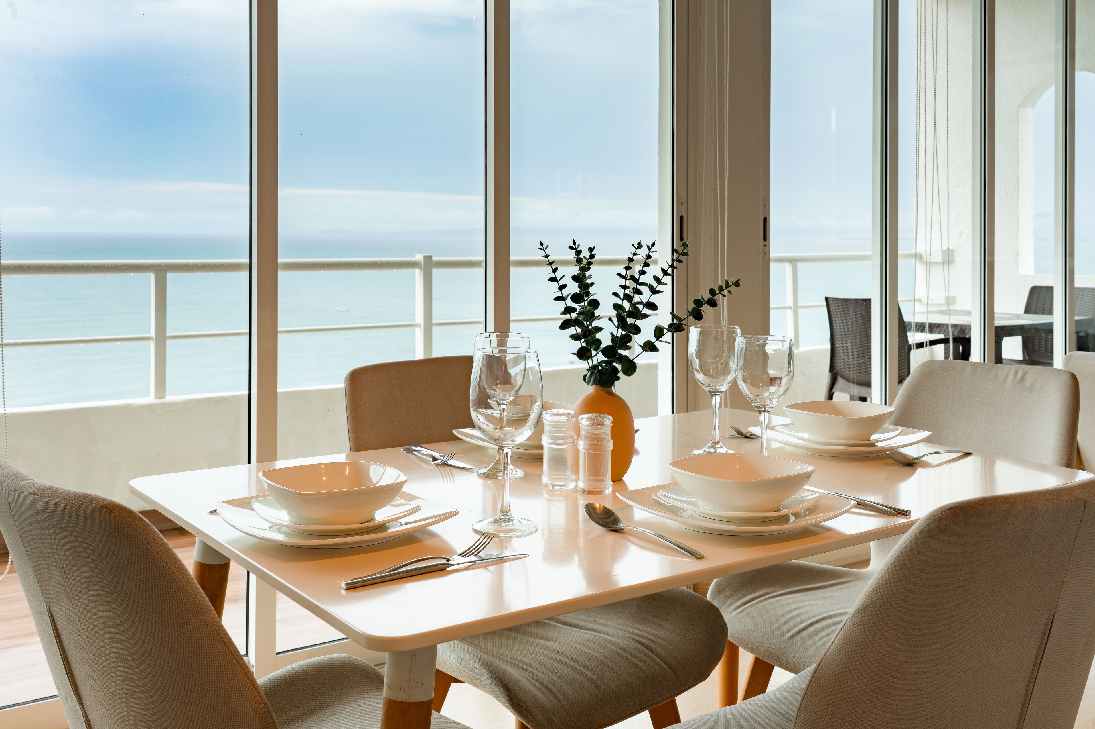 A dining table set for four with white plates, bowls, and cutlery, a vase with greenery, and a glass of water.