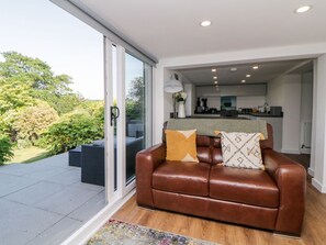A modern living room with a leather sofa, a glass door leading to a patio, and a kitchen in the background. - Riverview @ 1 Brunel View 1bed (Saltash)