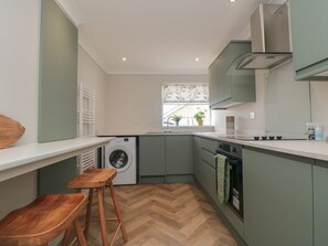 A compact kitchen with a washing machine, wooden stools, a window with a view, and a built-in oven. - 36 Chatsworth Close, Bridlington (Bridlington)