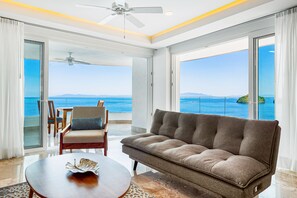A modern living room with a large sofa, a coffee table, and a dining area. The room has large windows offering a view of the sea. - Blue Horizon 901 | 1BR/2BA PV Ocean View, Pool, Gym (Puerto Vallarta)