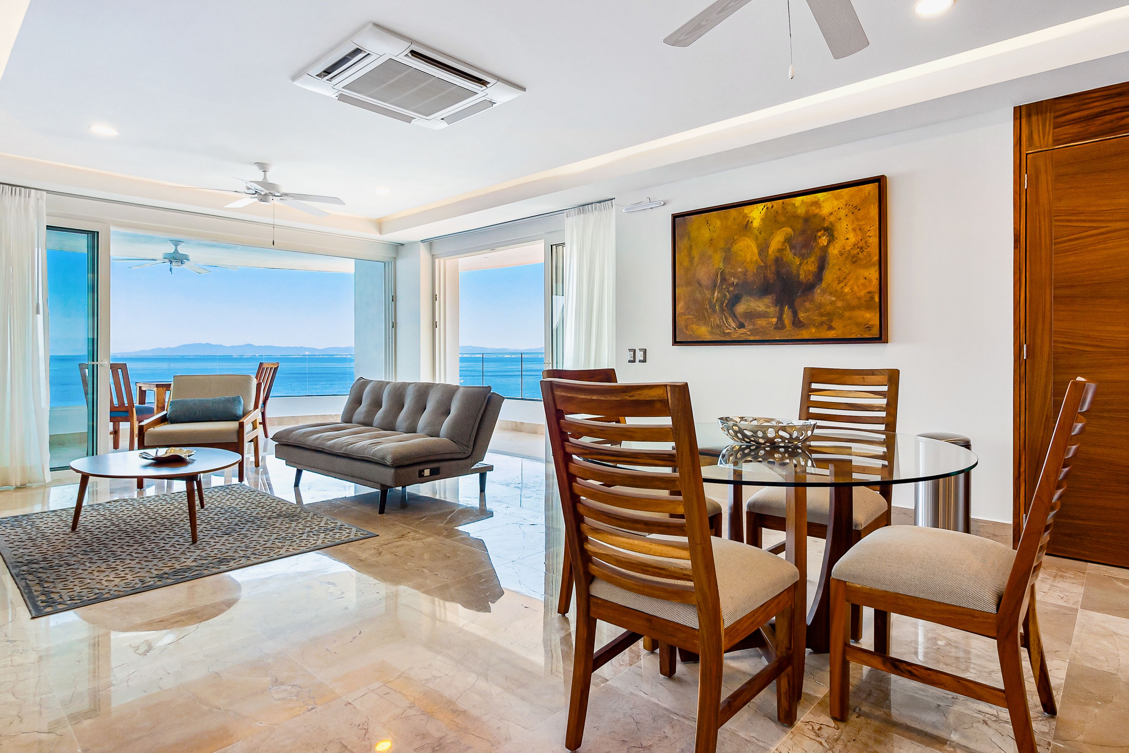 Una sala de estar moderna con una mesa de vidrio, sillas de madera, un sofá y vista al mar.