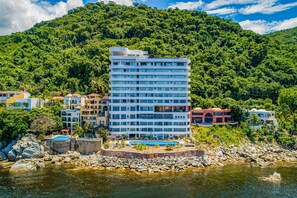 A multi-story hotel with a pool, surrounded by lush greenery and a rocky shoreline. - Blue Horizon 901 | 1BR/2BA PV Ocean View, Pool, Gym (Puerto Vallarta)