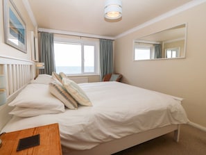 Ein Schlafzimmer mit Bett, Nachttisch, einem Fenster mit Vorhängen, einem Spiegel und Blick aufs Meer.