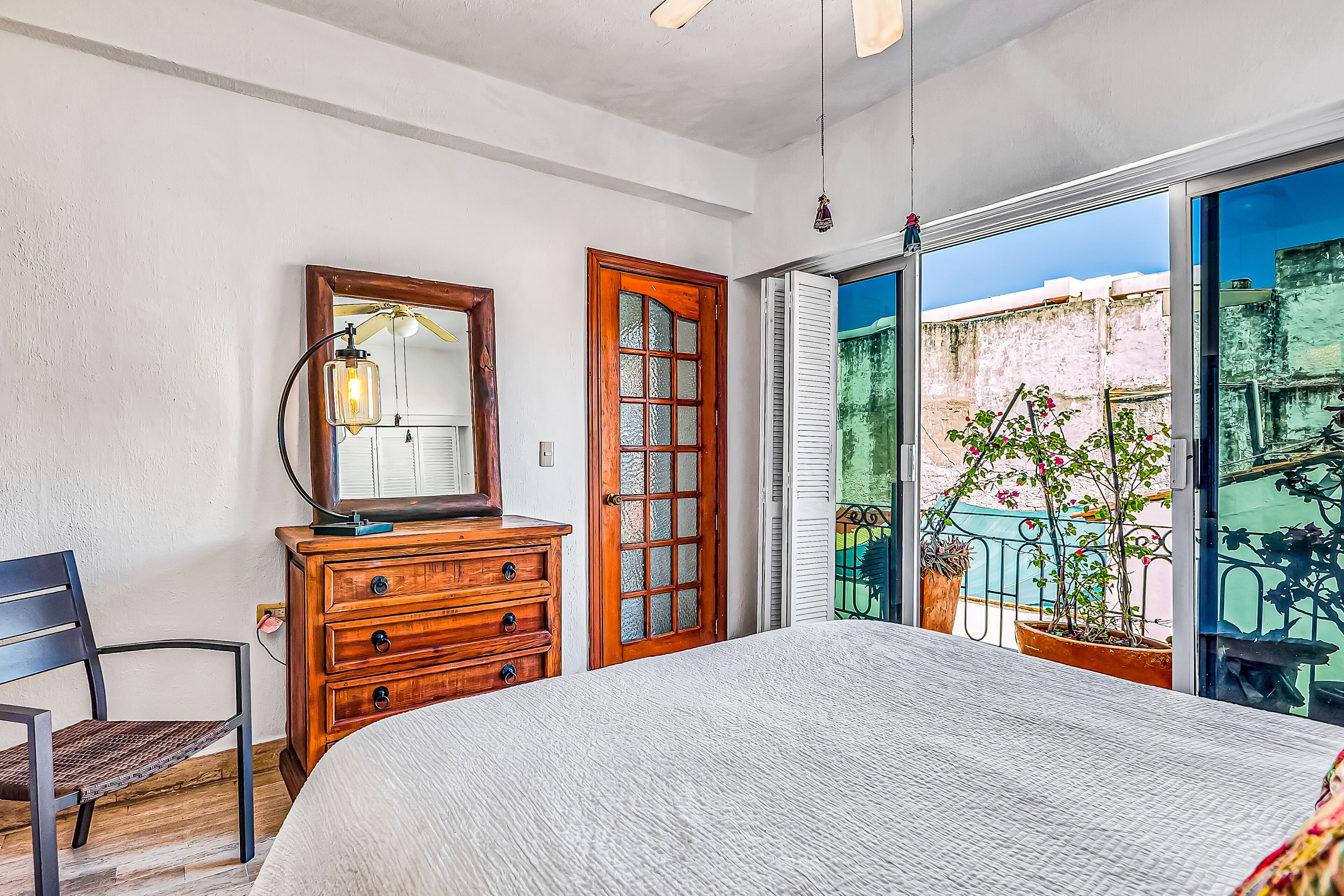 A bedroom with a large bed, wooden dresser, and a mirror. There is a balcony with potted plants visible through the door.