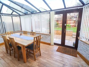 A conservatory with a wooden dining table and chairs, a glass door leading outside, and a brick wall. - Cherry Tree House, Penzance (Penzance)