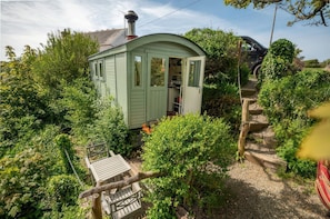 A small, light green wooden structure with a door and windows, surrounded by dense greenery and a gravel path. - The Shephard Hut, Solva (Haverfordwest)