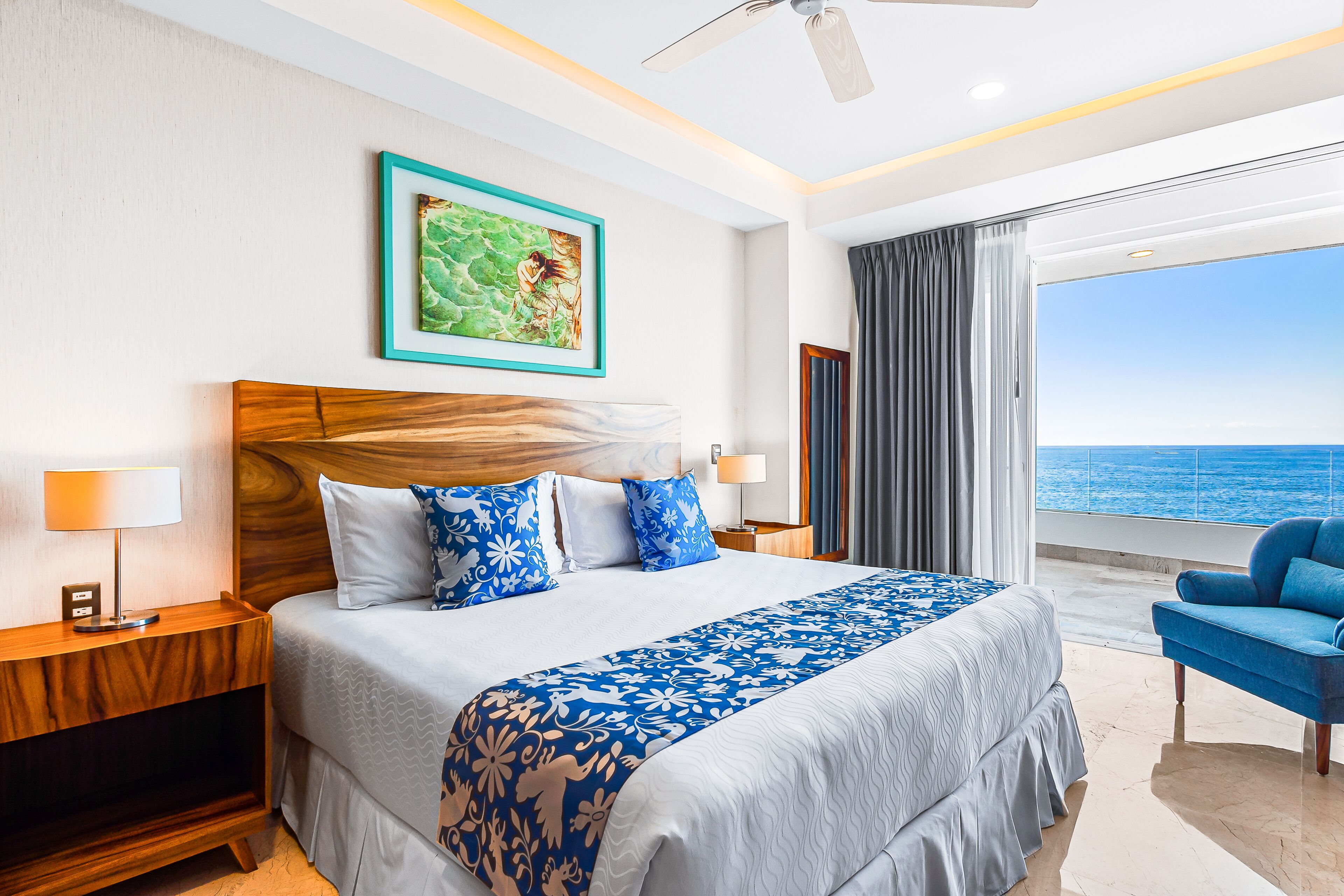 Una habitación de hotel con una cama grande, vista al océano, ventilador de techo y una silla azul.