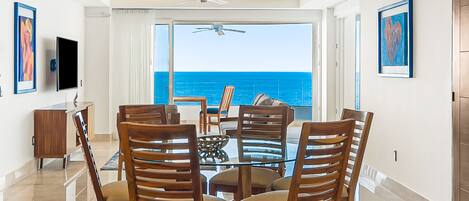 A dining area with a glass table and wooden chairs, a ceiling fan, and a view of the ocean.