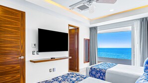 Une chambre d’hôtel équipée d’une télévision, de deux lits avec une literie à motifs floraux bleus, d’une armoire en bois et d’un balcon avec vue sur la mer.