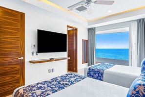 Ein Hotelzimmer mit Fernseher, zwei Betten mit blau-blumigem Bettbezug, einem Holzschrank und einem Balkon mit Meerblick.