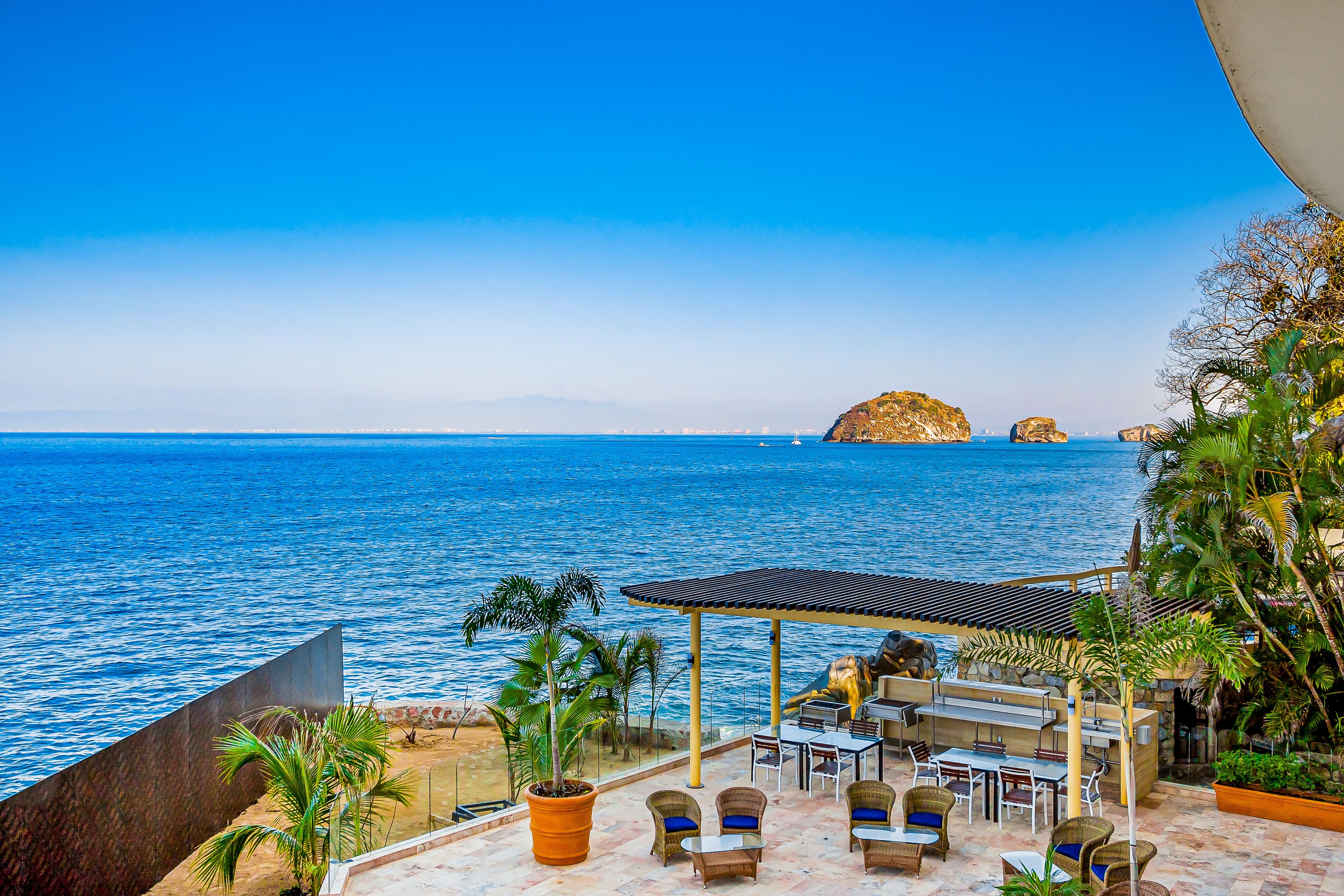 Eine Terrasse mit Sitzgelegenheiten im Freien, Blick auf das Meer und Palmen.