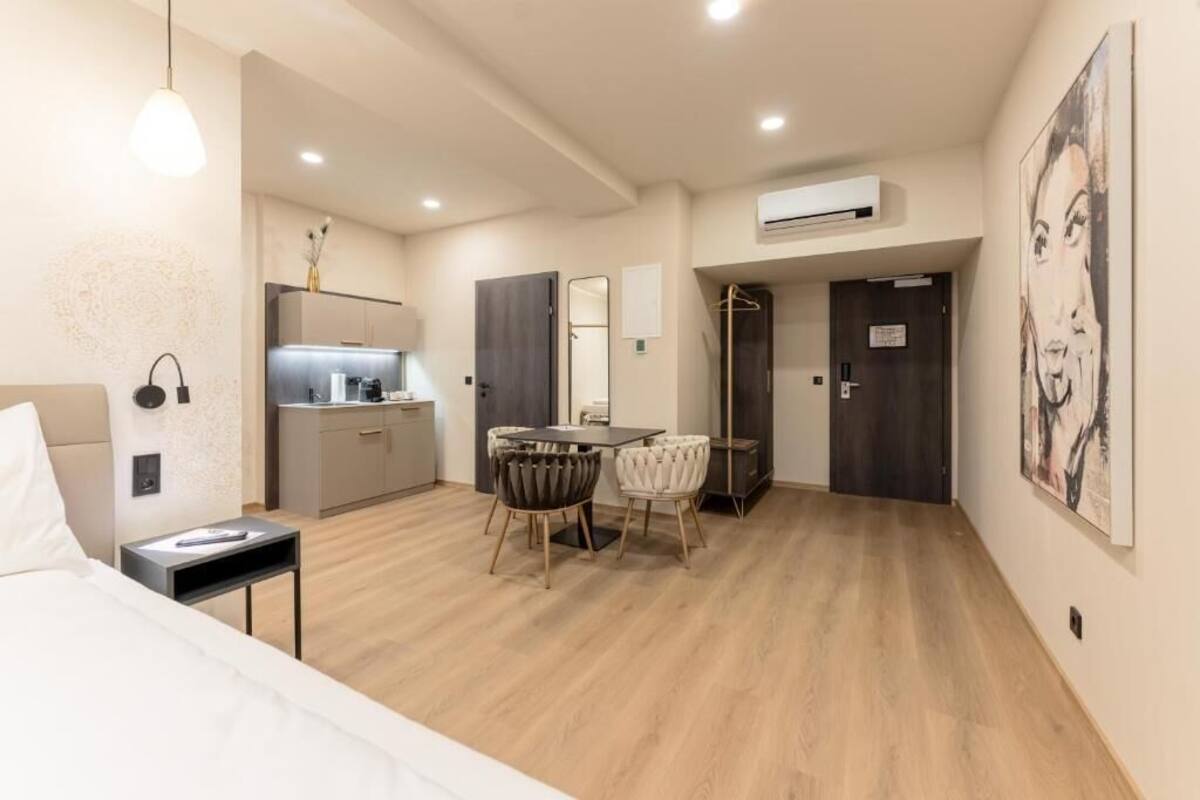 A modern hotel room with a bed, bedside table, desk, chairs, kitchenette, and artwork on the walls.