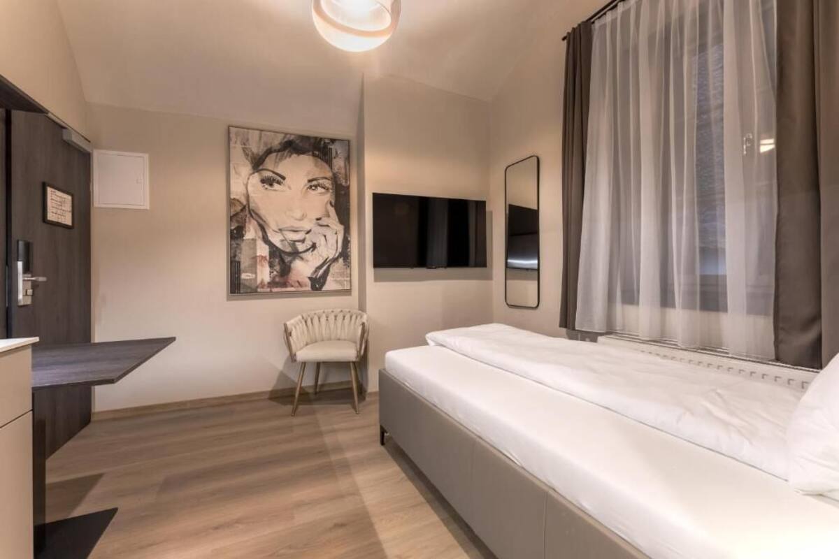 A modern hotel room with a bed, a chair, a desk, a TV, and a large painting on the wall.