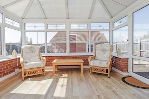 A conservatory with two wicker chairs, a wooden bench, and large windows offering a view of a red-brick building. - Echo Beach, Seasalter (Whitstable)