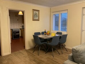 A dining area with a table, chairs, a kitchen in the background, and a window with blinds.