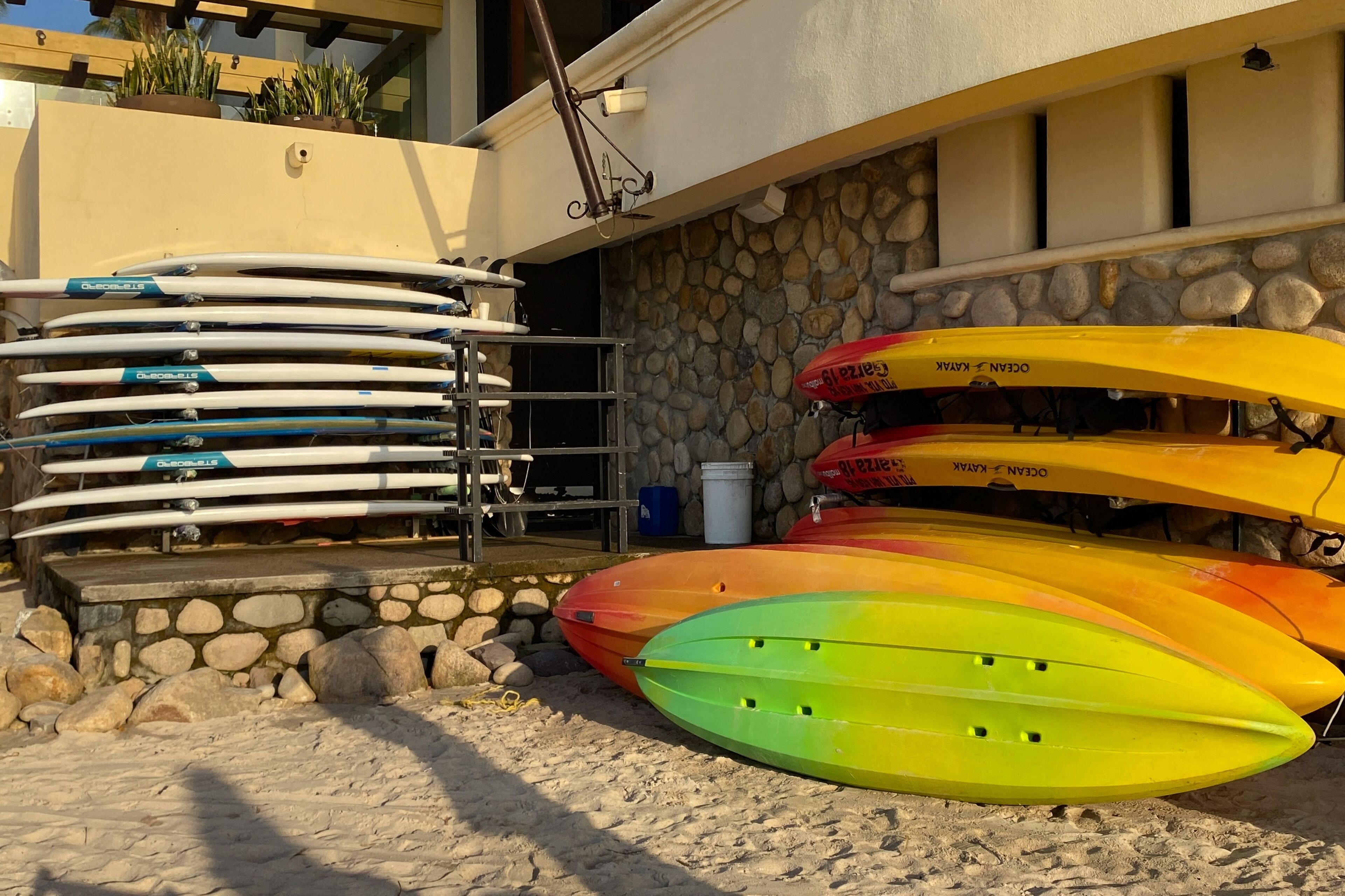 A collection of kayaks and paddleboards stacked against a building with a stone wall and a metal rack.