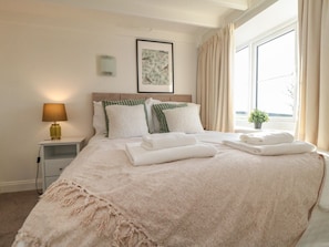 A neatly made bed with pillows and folded towels, a bedside lamp, a framed picture on the wall, and a window with curtains.