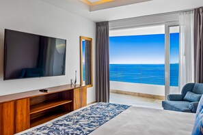 Ein Hotelzimmer mit einem großen Flachbildfernseher, einer Holzunterhaltungseinrichtung, einem Bett mit gemustertem Überwurf, einem blauen Sofa und einem Balkon mit Meerblick.