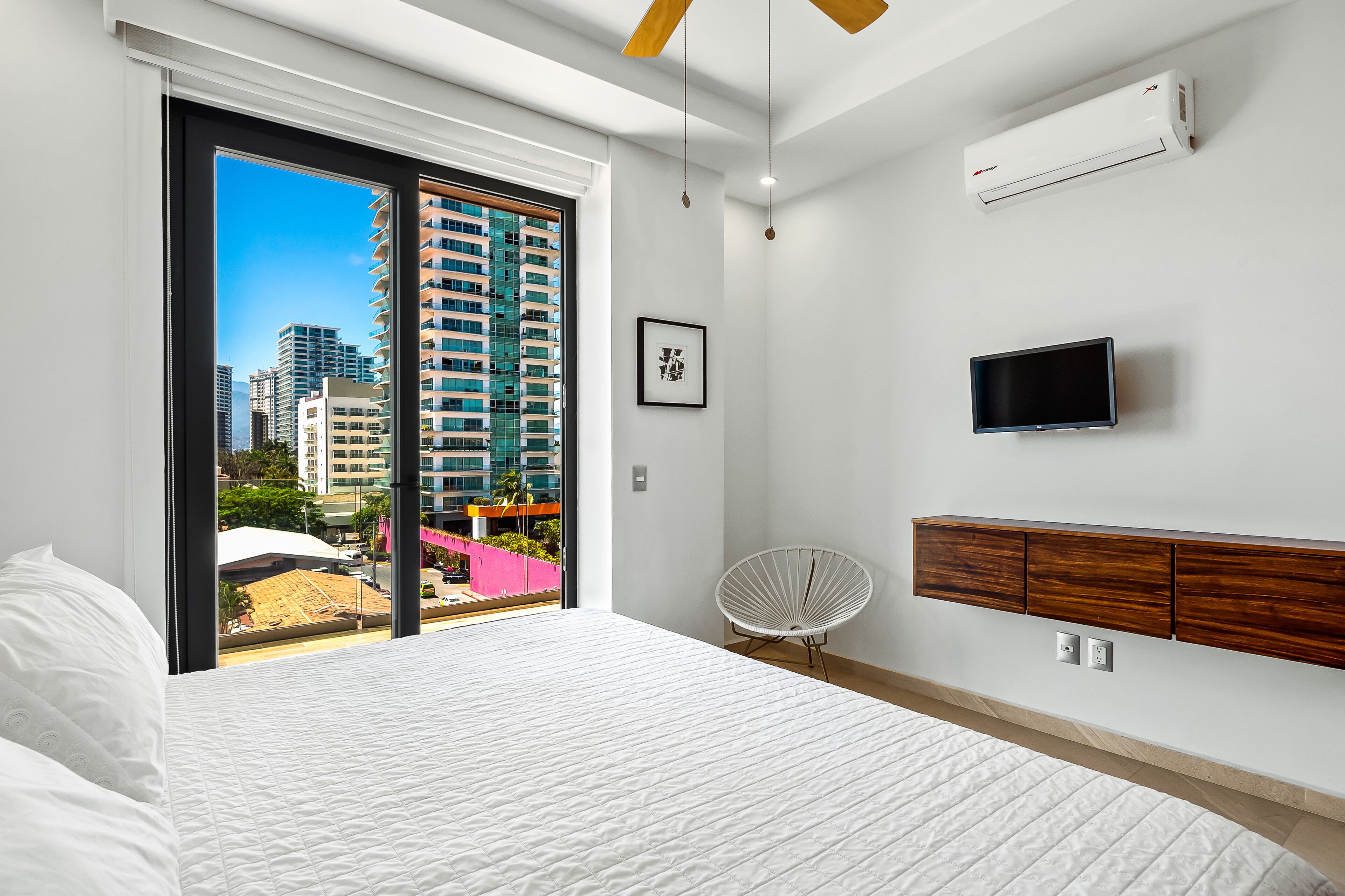 A modern hotel room with a large bed, a city view through the window, a mounted TV, and a ceiling fan.