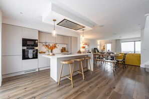 A modern kitchen with a central island, wooden stools, and a dining area with a yellow sofa. - Porth Beach Villa 9 Longshore, Newquay (Newquay)