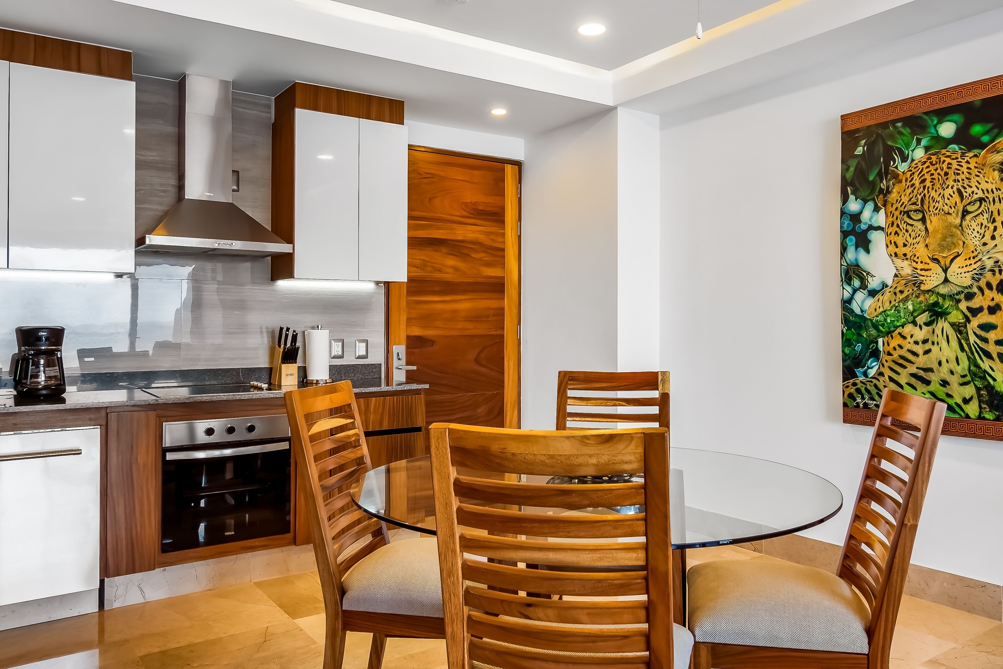 A modern kitchen with wooden cabinets, a built-in oven, and a glass dining table with wooden chairs.