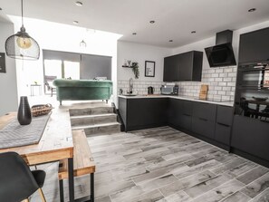 A modern kitchen with dark cabinetry, a wooden dining table, and a green sofa. - Charming Ty Coch in Ruthin (Ruthin)
