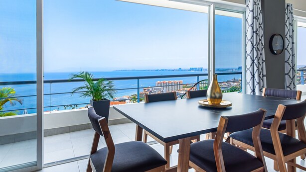 A dining area with a large table and chairs, a vase, and a view of the ocean and cityscape. - Dog-friendly ocean view penthouse & studio with a full kitchen & kitchenette (Puerto Vallarta)