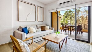 Un salon moderne avec un canapé, une table basse et des fauteuils en osier. Une porte-fenêtre coulissante donne accès à une terrasse extérieure équipée d’une table et de chaises.