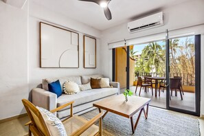 Een moderne woonkamer met een bank, salontafel en rieten stoelen. Er is een glazen schuifdeur die leidt naar een terras met een tafel en stoelen.