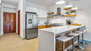 Une cuisine moderne avec un réfrigérateur en acier inoxydable, une cuisinière à gaz, des armoires en bois et un comptoir blanc. On y trouve trois tabourets de bar et des luminaires suspendus au-dessus du comptoir.