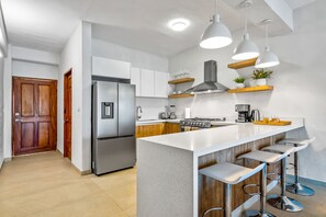 Een moderne keuken met een roestvrijstalen koelkast, een gasfornuis, houten kastjes en een wit aanrecht. Drie barkrukken en hanglampen hangen boven het aanrecht.