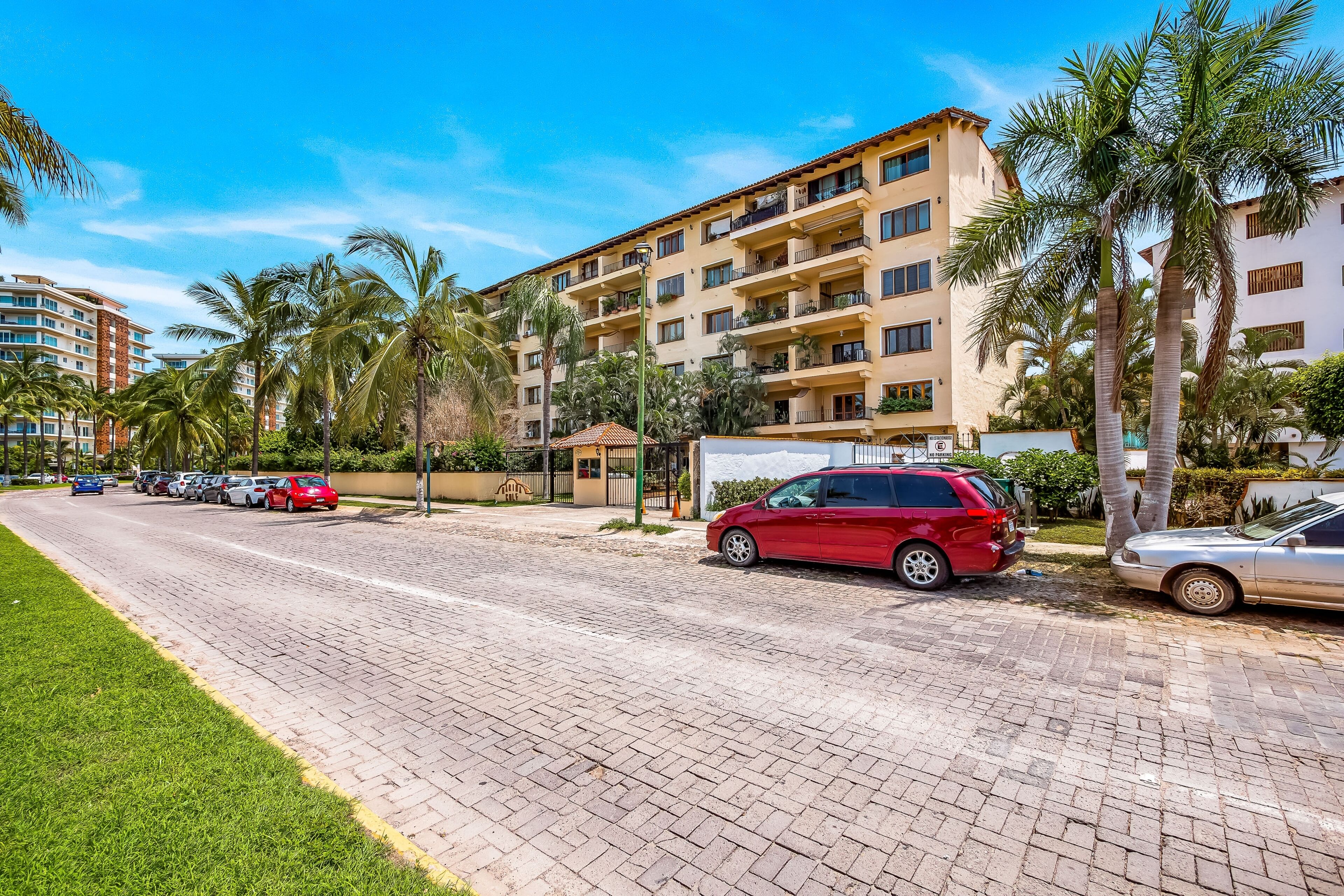A multi-story hotel with balconies, palm trees, and a paved driveway with parked cars.