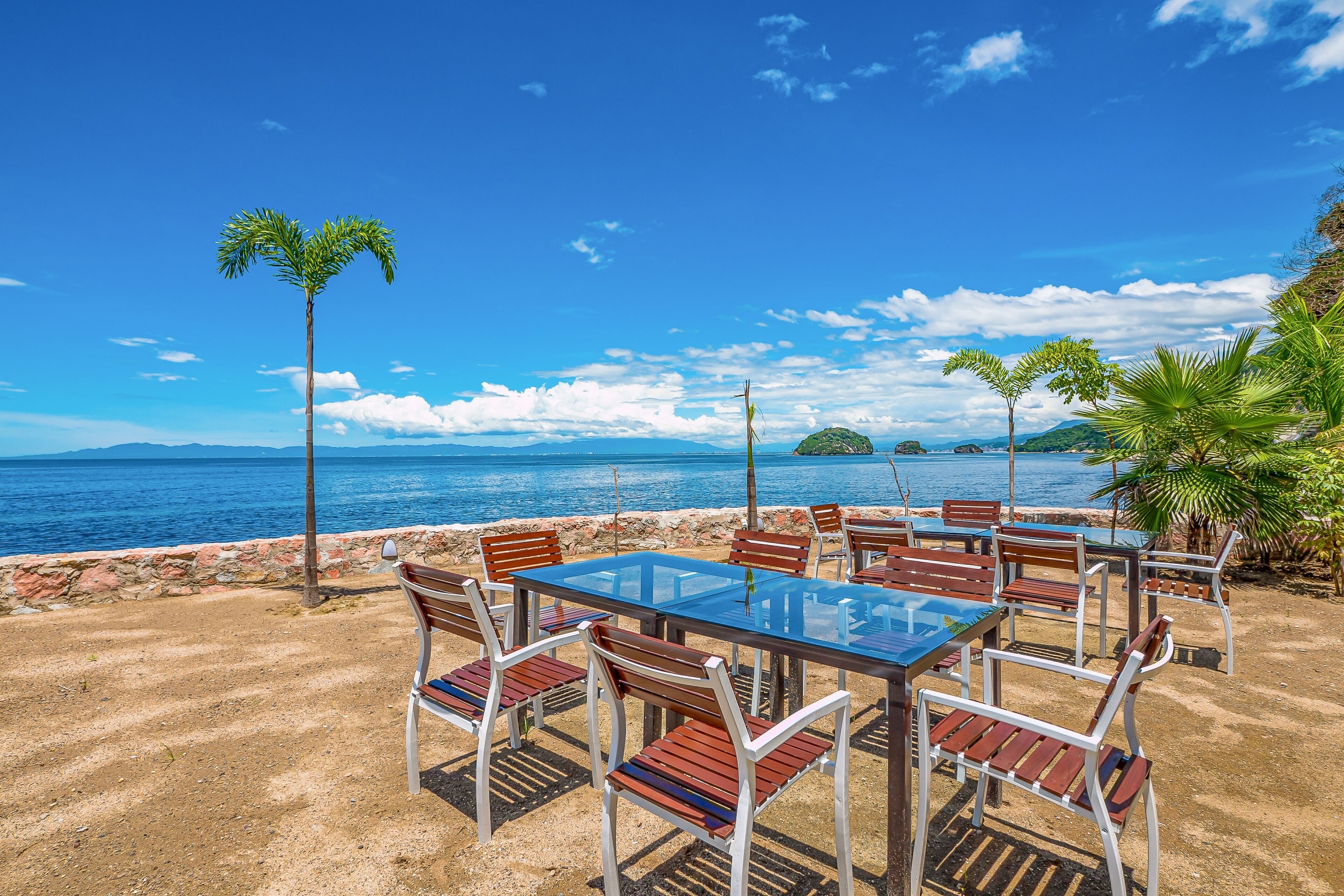 A beachside dining area with a glass table and chairs, palm trees, and a view of the sea.