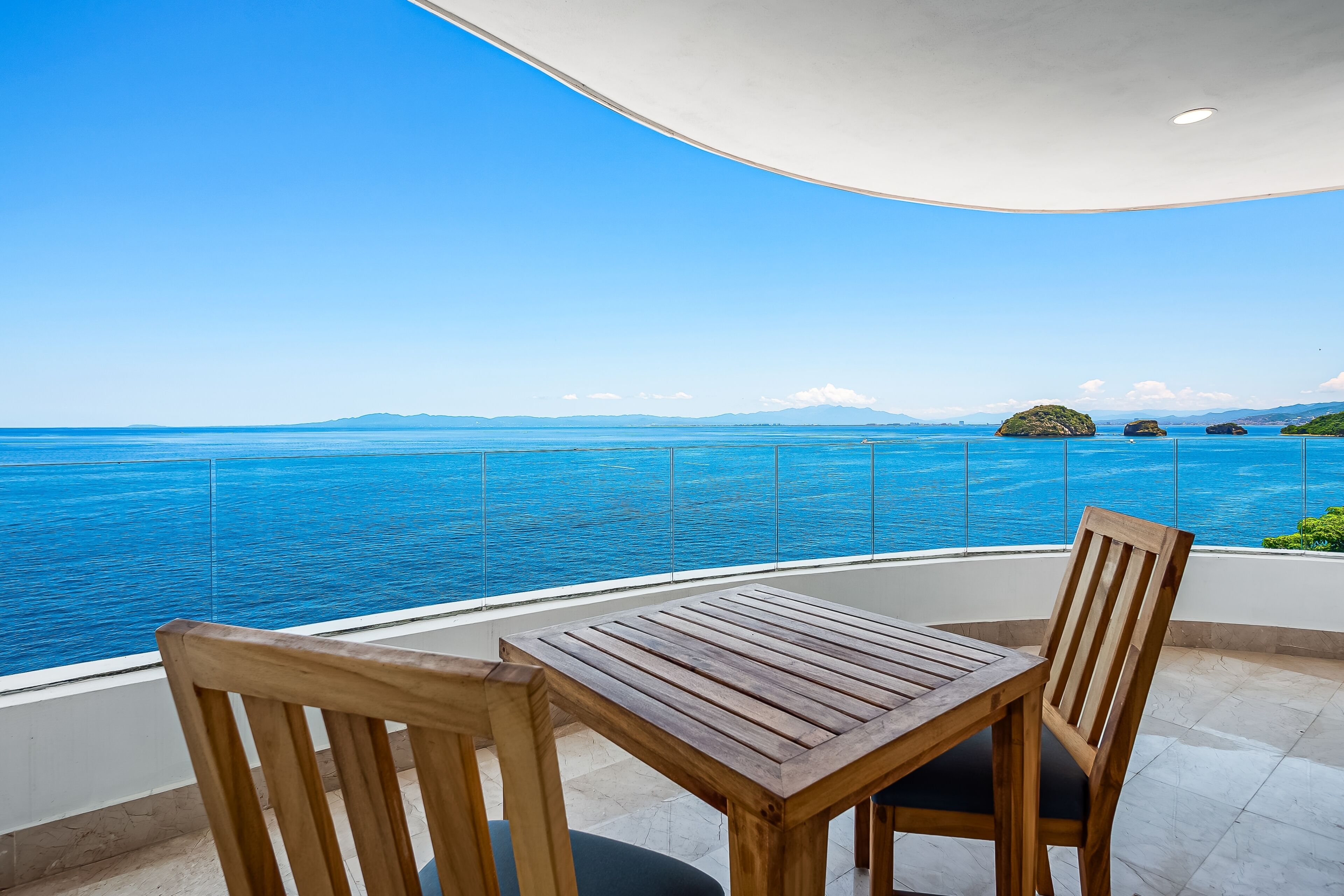 A wooden table and chairs on a balcony with a clear view of the sea and distant islands.