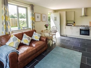 A living room with a brown leather sofa, a blue rug, a dining table with chairs, and a kitchen with white cabinets and a stove. - The Lodge, Lower Trefedw, Pandy, South Wales (Abergavenny)